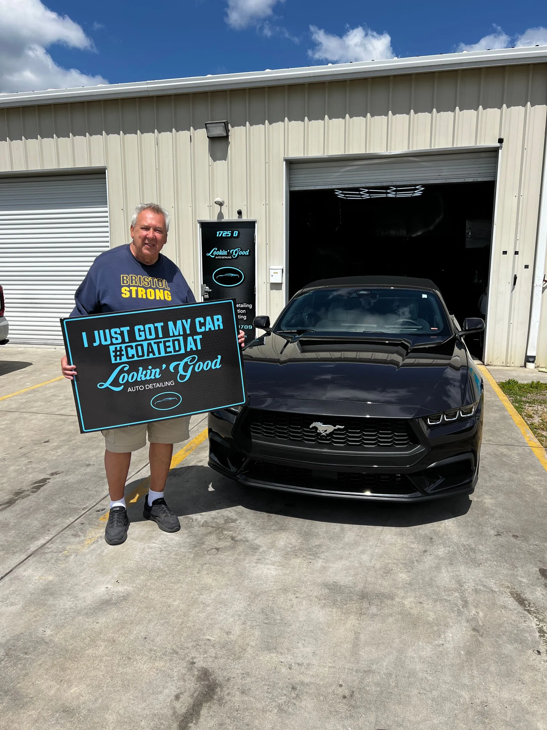 Client holding a #COATED sign beside a coated Ford Mustang at the shop